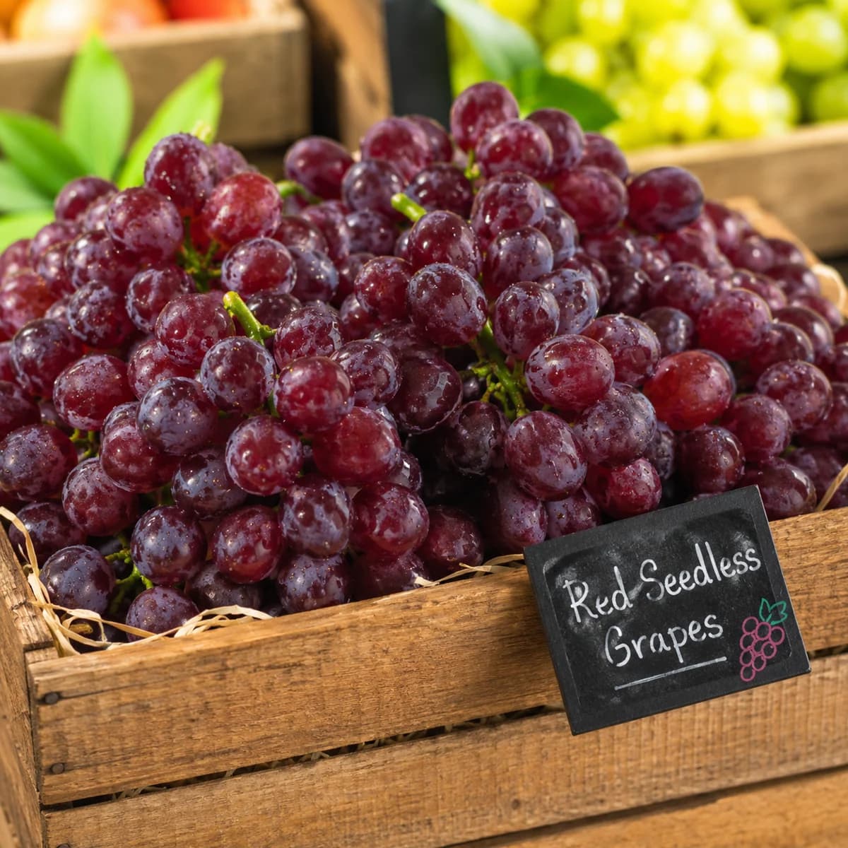Red grapes arranged in a wooden market crate beside a small English sign reading Red Seedless Grapes